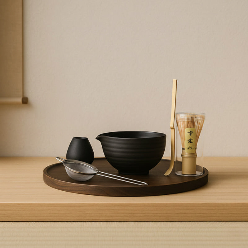 Tea-making set with black bowl, bamboo whisk, and other tools on a wooden tray against a beige wall.