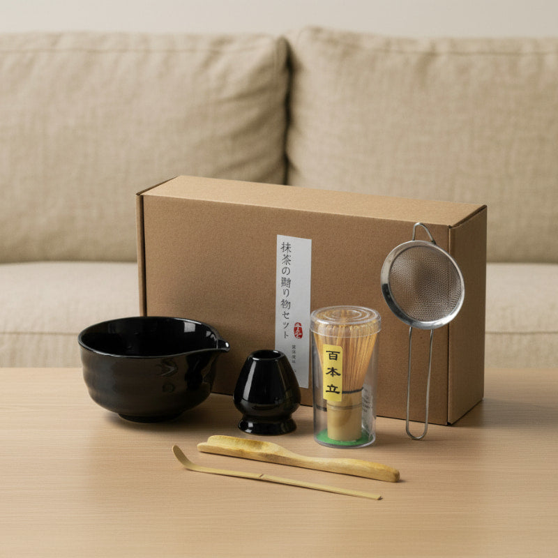 classic tea set design with a black bowl, black scoop, green container, wooden sticks, and metal strainer in front of a cardboard box.