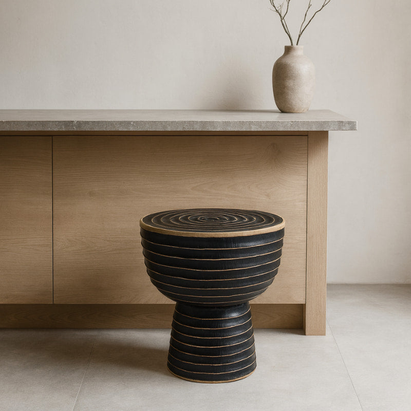 Black textured wabi sabi handmade kitchen stool in front of a wooden cabinet with a vase on top.