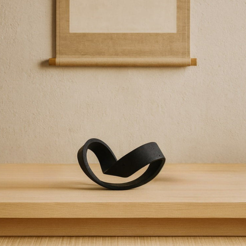 wabi sabi house decor sculpture between two beige bowls on a wooden surface with a beige wall and scroll in the background.