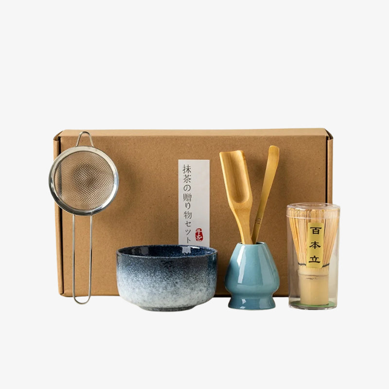 Tea-making set with a bowl, glass, and whisk tool in front of a cardboard box on a white background