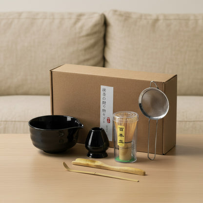 classic tea set design​ with a black bowl, black scoop, green container, wooden sticks, and metal strainer in front of a cardboard box.