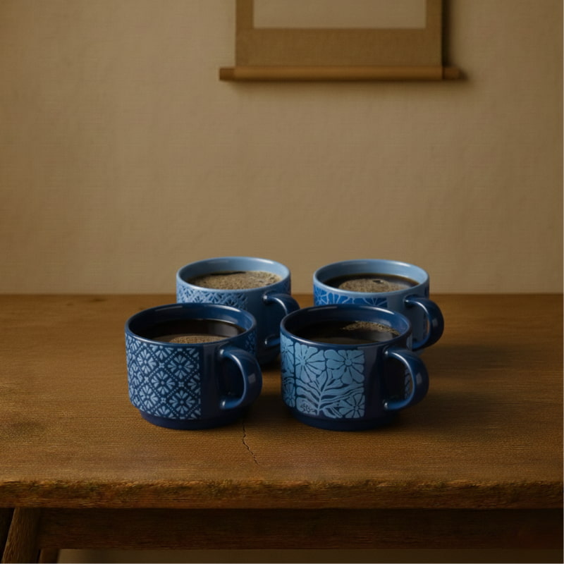 Four blue designer coffee mugs​ with different patterns on a wooden table against a beige wall.