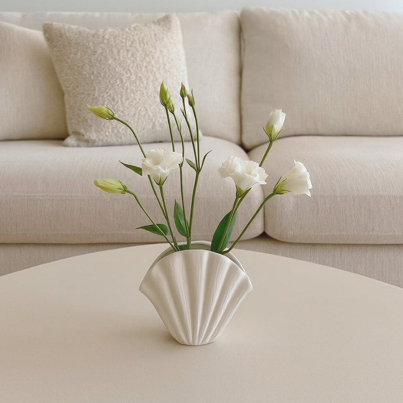 Shell-shaped wabi sabi style pot with white flowers on a coffee table in front of a beige sofa.
